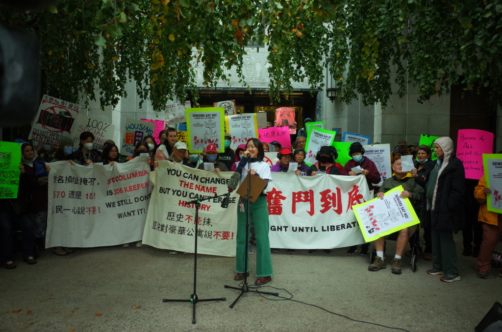 Community Activist Jade Ho speaking at a rally in front of City Hall on Monday October 20, 2025. Chinatown community members gathered behind Jade hold banners and signs with slogans including "奮鬥到底 Fight Until Liberation", "You can change the name but you can't change the history", and more. Photo Credit: Alger Liang. Photo Source: Alger Liang.
