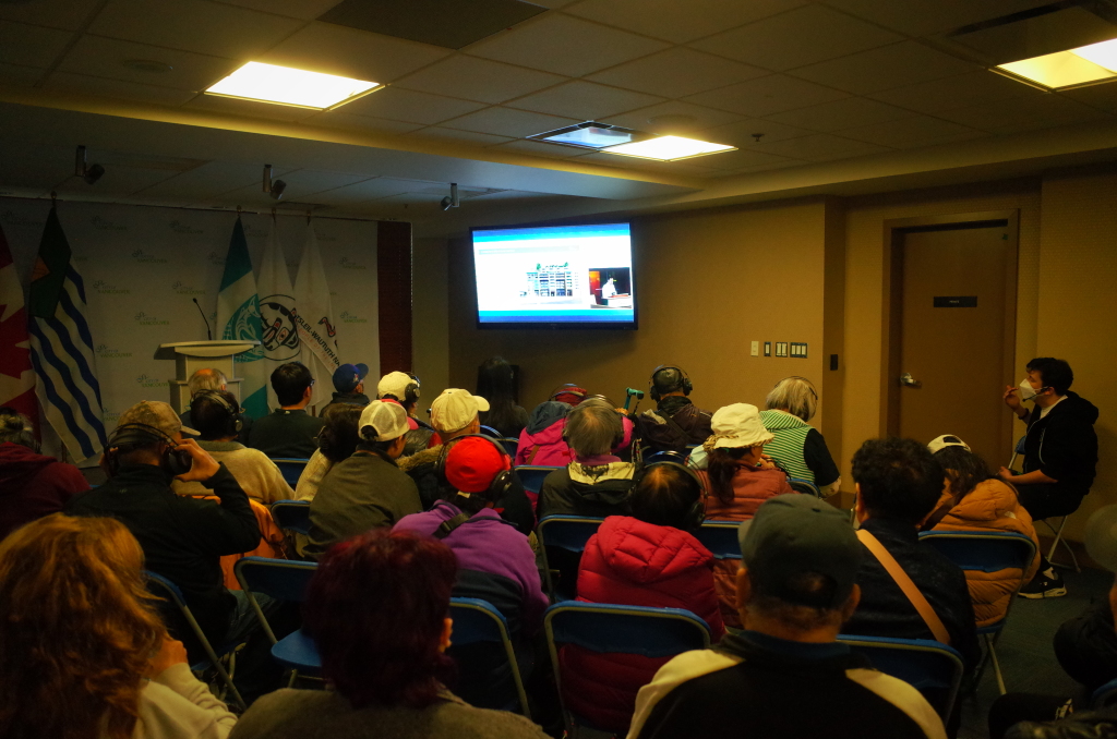Chinatown community members gathered in the Joe Wai room at City Hall for the 105 Keefer (570 Columbia) Development Permit Board hearing on Monday October 20, 2025. The elders, activists, and allies are seated on blue folding plastic chairs in front of a wall-mounted TV streaming the hearing. Photo Credit: Alger Liang. Photo Source: Alger Liang.